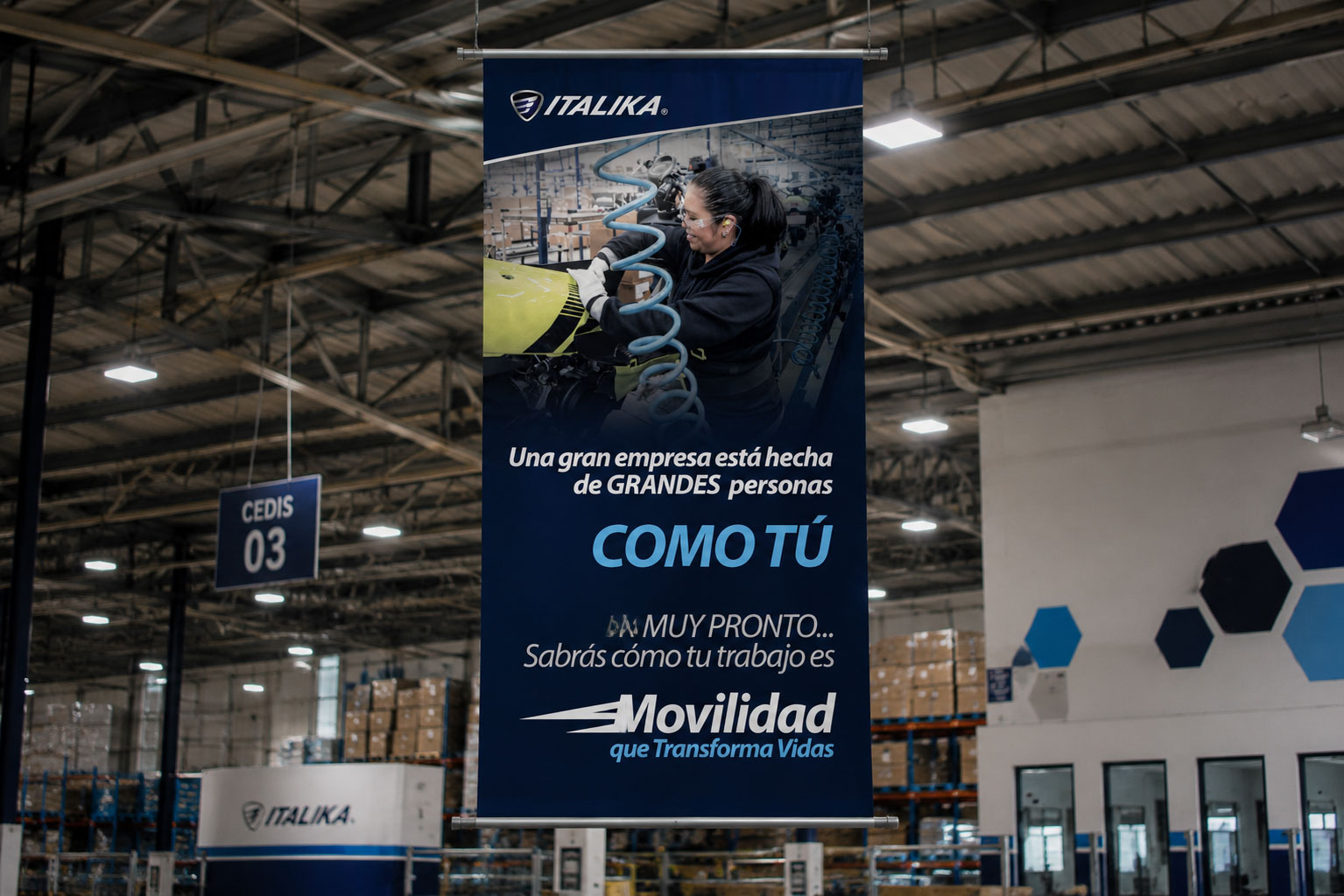 Banner colgante de ITALIKA en un centro de distribución con una trabajadora y el mensaje "Una gran empresa está hecha de grandes personas como tú"