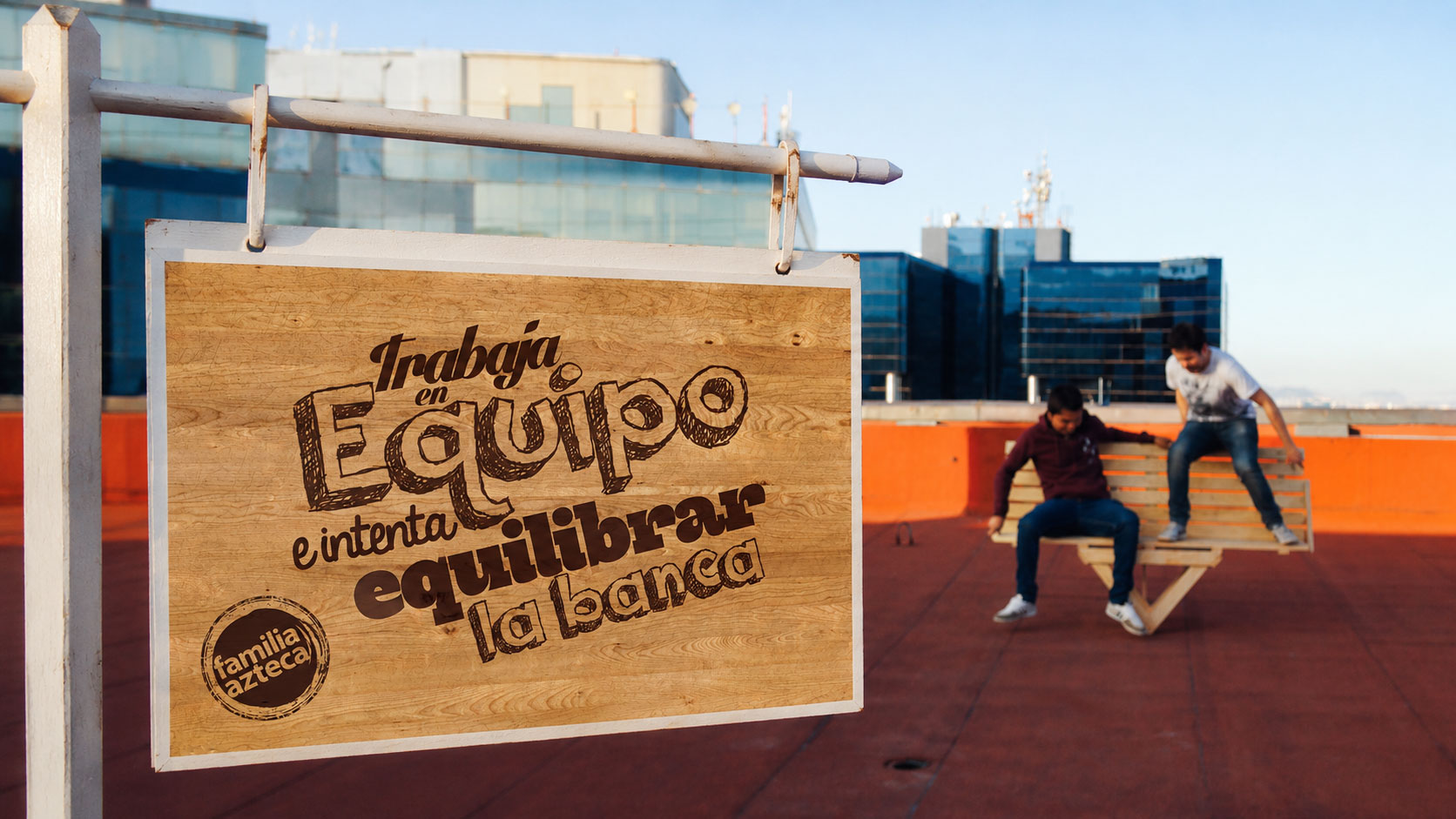 Letrero de madera «Trabaja en equipo e intenta equilibrar la banca» junto a dos personas sentadas en los extremos de la banca en una azotea