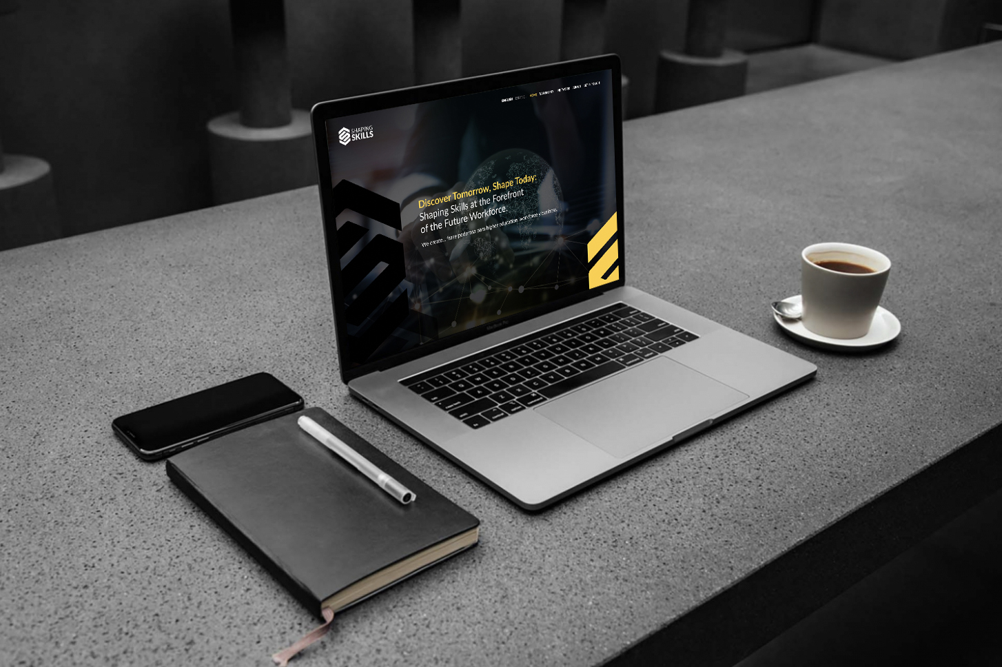 Laptop sobre un escritorio de concreto con el sitio web de Shaping Skills, junto a una libreta, un teléfono y una taza de café