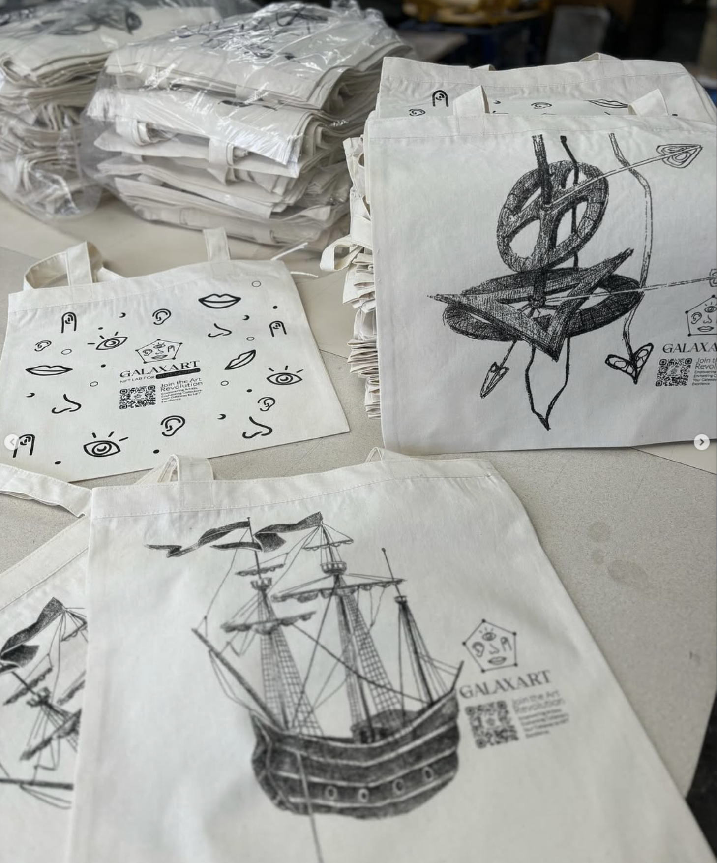 Pilas de bolsas tote de Galaxart impresas sobre una mesa, con el dibujo de un velero y un patrón de rostros